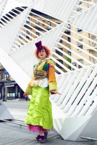 A woman in a highly detailed, avant-garde outfit featuring a massive floral headpiece and a red silk robe, posing in front of ornate bronze doors.
