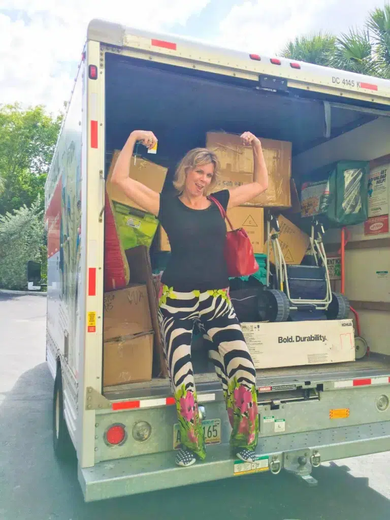 A smiling woman in colorful pants and a black shirt poses playfully, flexing her arms while sitting on the edge of an open moving truck filled with boxes&mdash;perhaps ready to call on Miami interior designers for her new space.